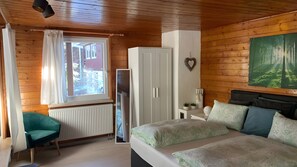 Une pièce aux murs lambrissés, avec un lit, une armoire blanche, un radiateur, une fenêtre avec des rideaux et un tableau accroché au mur.