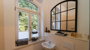 A bathroom with a window, a sink, a mirror, and a shelf with toiletries.