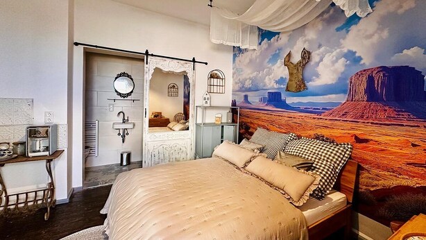 A bedroom with a large bed, a dresser, a vanity, and a mural of a desert landscape. - Little Honeymoon Suite (Penig)