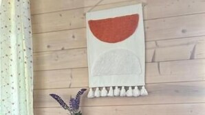 Una habitación con paredes de madera, alfombra roja estampada, dos sillas de madera, una toalla blanca con flecos y un jarrón con flores moradas.