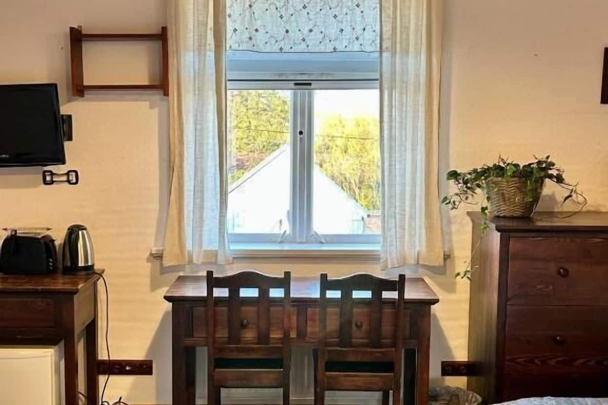 A room with a wooden bench, a bed, a window with curtains, a television, and a plant.