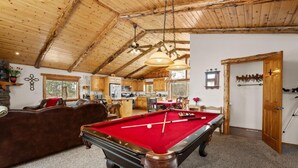 Une table de billard dans une pièce avec des poutres en bois au plafond, un canapé et une cuisine en arrière-plan.