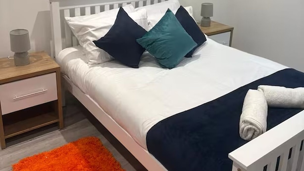 Een bed met witte beddengoed, donkere kussens en een blauwe dekens. Er is een wit hoofdbord, een nachtkastje met een lamp, en een kleine tafel met een lamp. Op de vloer ligt een oranje vloerkleed.