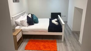 Een eenpersoonsbed met een wit hoofdbord, een nachtkastje, een feloranje vloerkleed en een schilderij aan de muur.