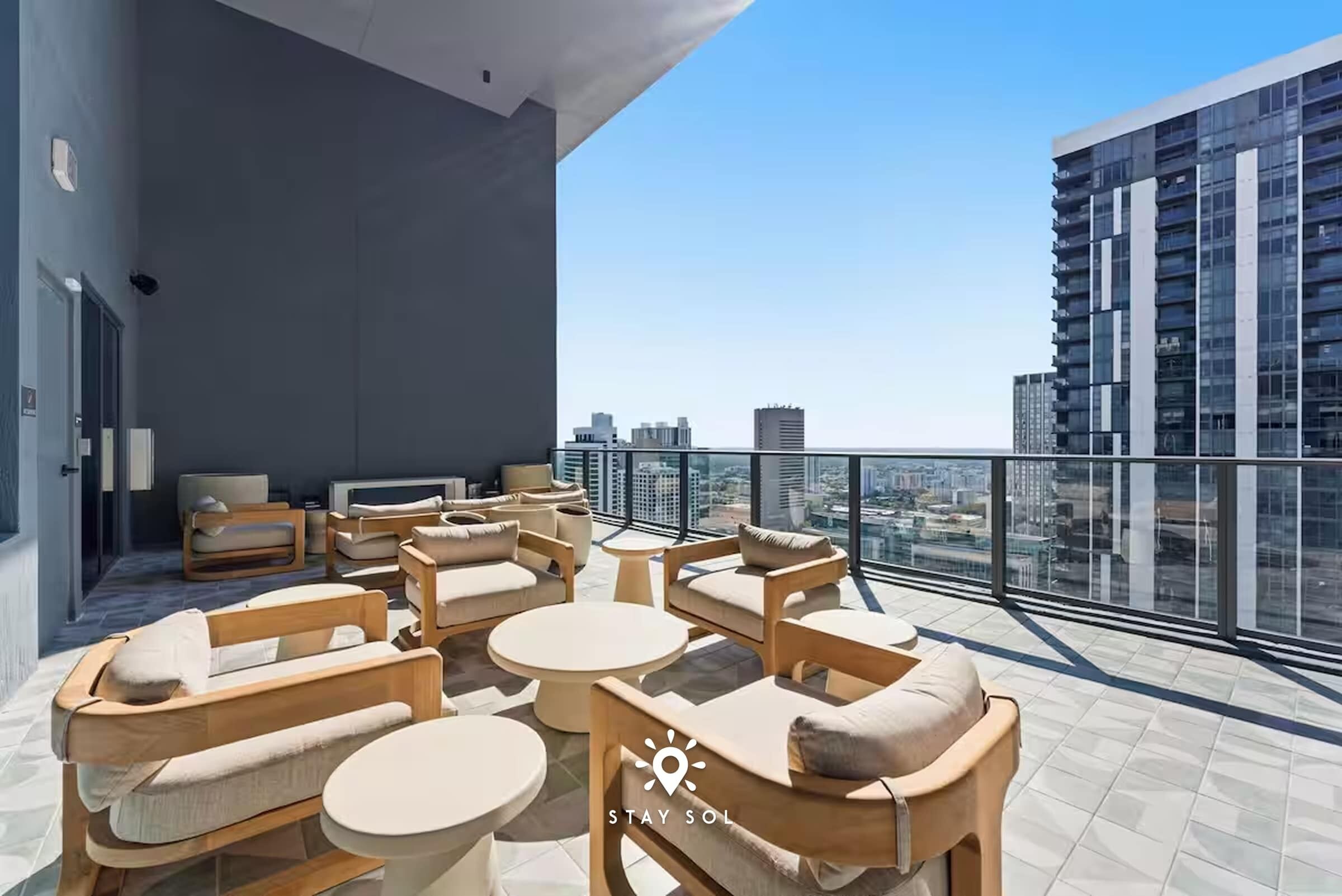 Un salón en la azotea con mesas redondas y sillas de madera, vistas de la ciudad y un edificio moderno al fondo.