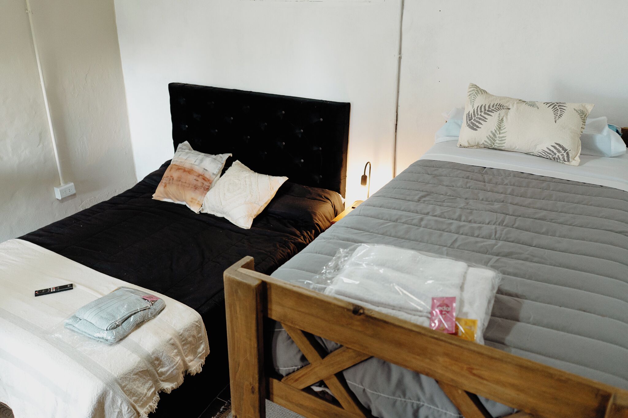 Une chambre avec deux lits, dont l’un a une tête de lit noire et l’autre un cadre en bois. Une table de chevet avec une lampe se trouve à côté du lit de droite.