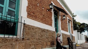 A person sitting on a bench outside a building with a stone wall, a metal fence, and a street lamp. - Serrano Lodging in the Sierras of Córdoba (Achiras)