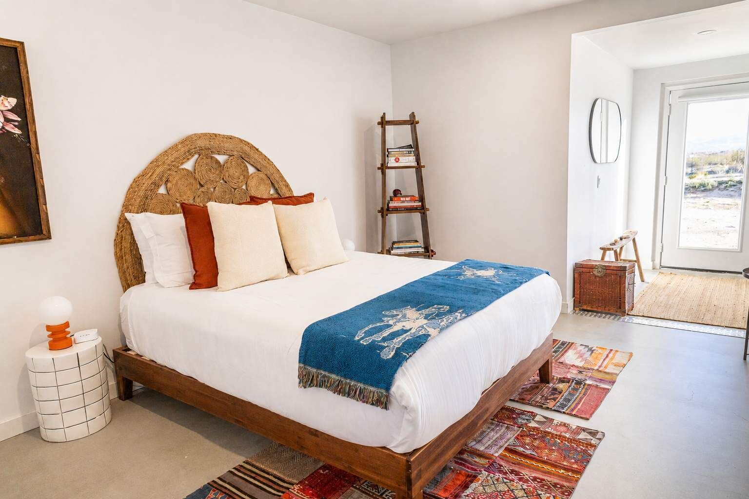 A bedroom with a bed, a bedside table, a nightstand, a wooden ladder, a suitcase, and a view of the outside.