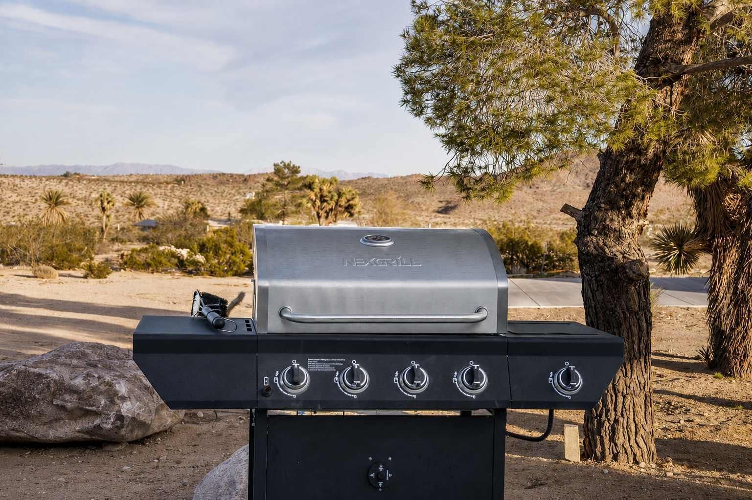 A portable grill with four burners and a lid, set outdoors with a desert landscape in the background.