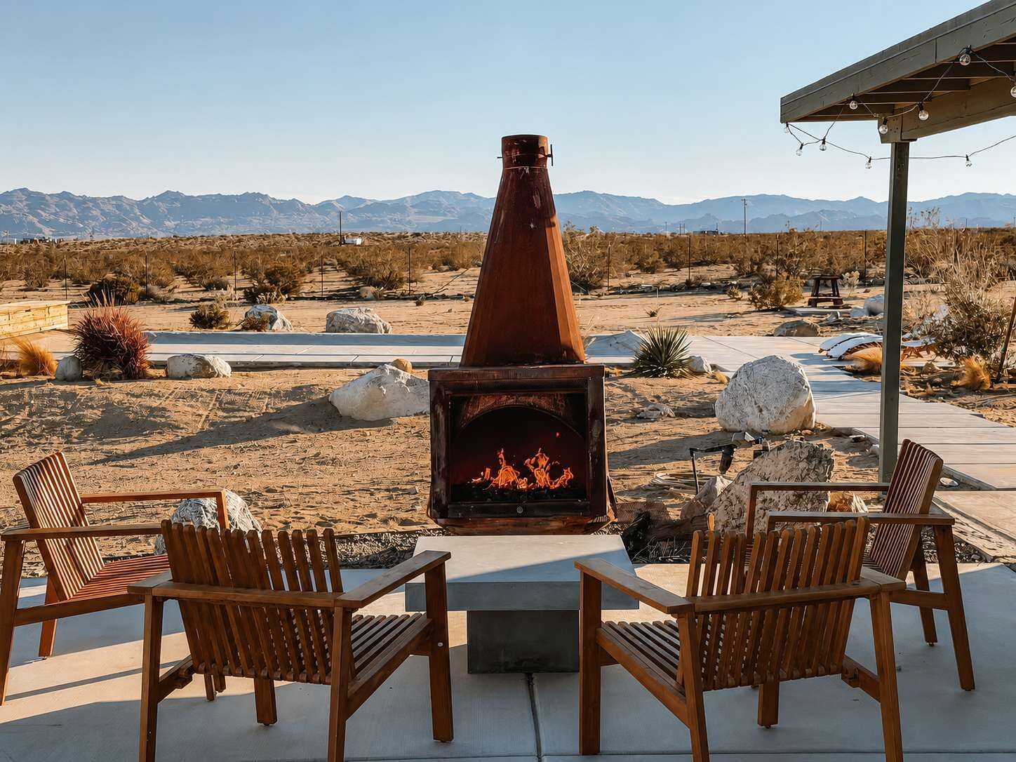 Un patio al aire libre con chimenea de leña, rodeado de sillas de madera, frente a un paisaje desértico con montañas al fondo.