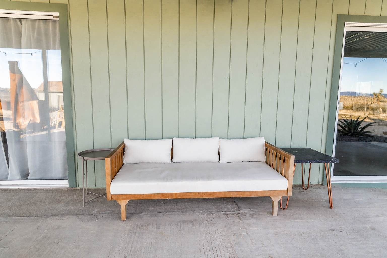 A wooden sofa with cushions against a green wall, a small round table beside it, and a view of a desert landscape through a glass door.