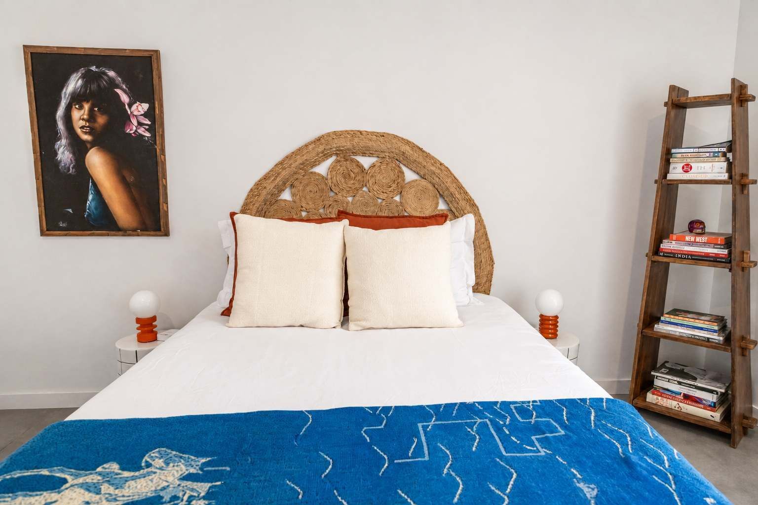 A bed with a woven headboard, two pillows, a blue patterned bedspread, a wooden bookshelf with books, and a framed picture on the wall.