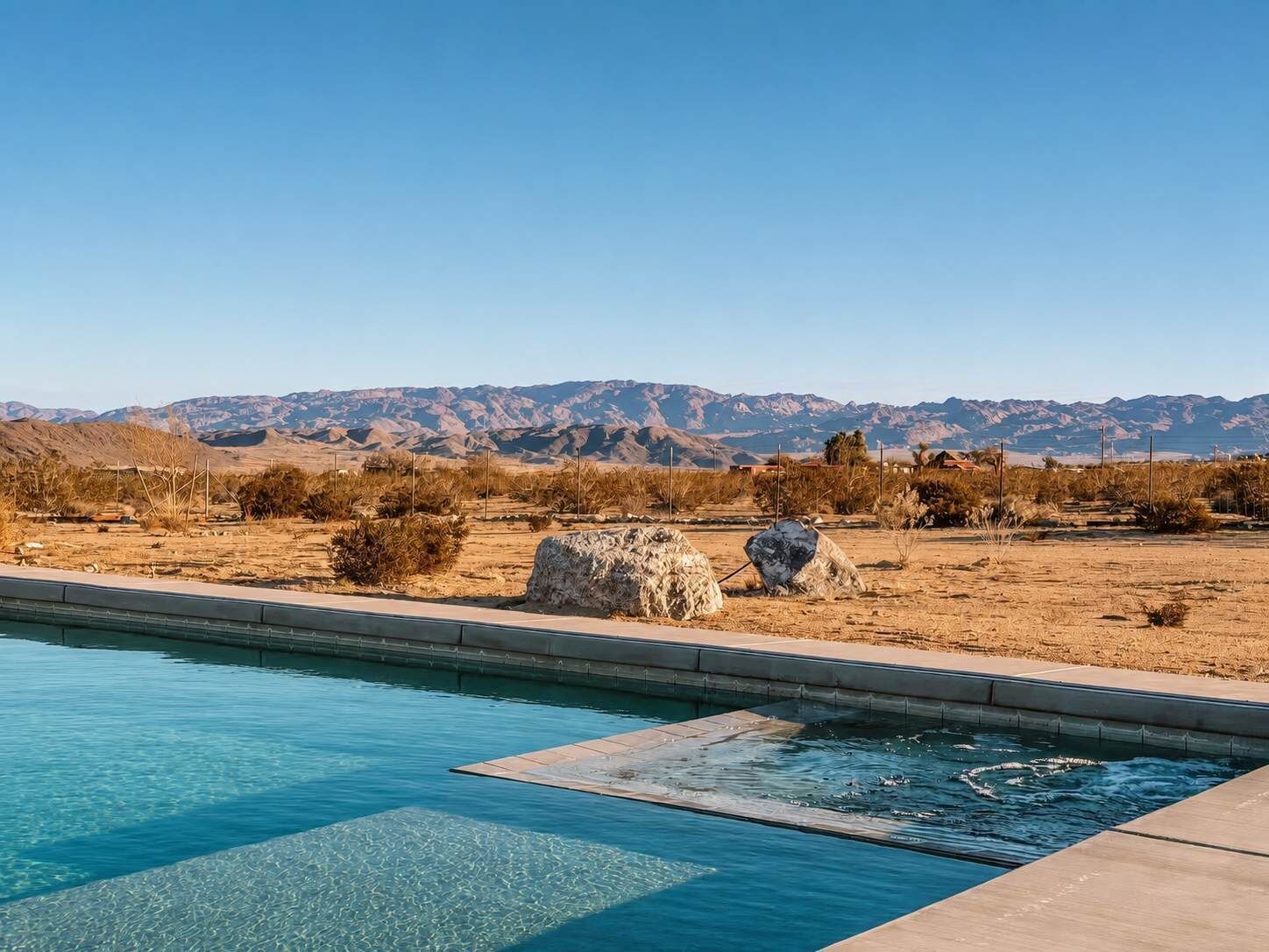 A swimming pool with clear water, surrounded by dry vegetation and mountains in the background.