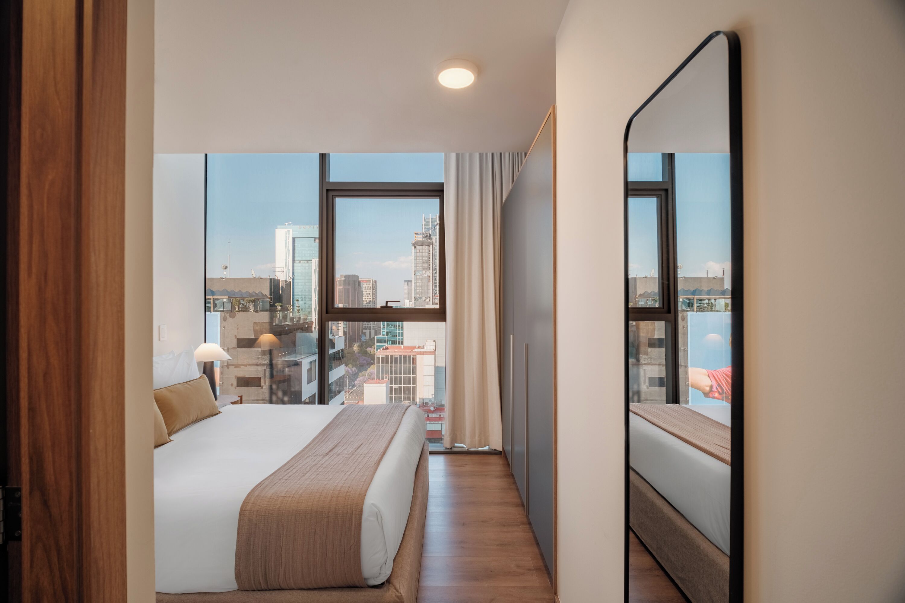 Ein Hotelzimmer mit großem Fenster, einem Bett, einem Nachttisch und einem Spiegel.
