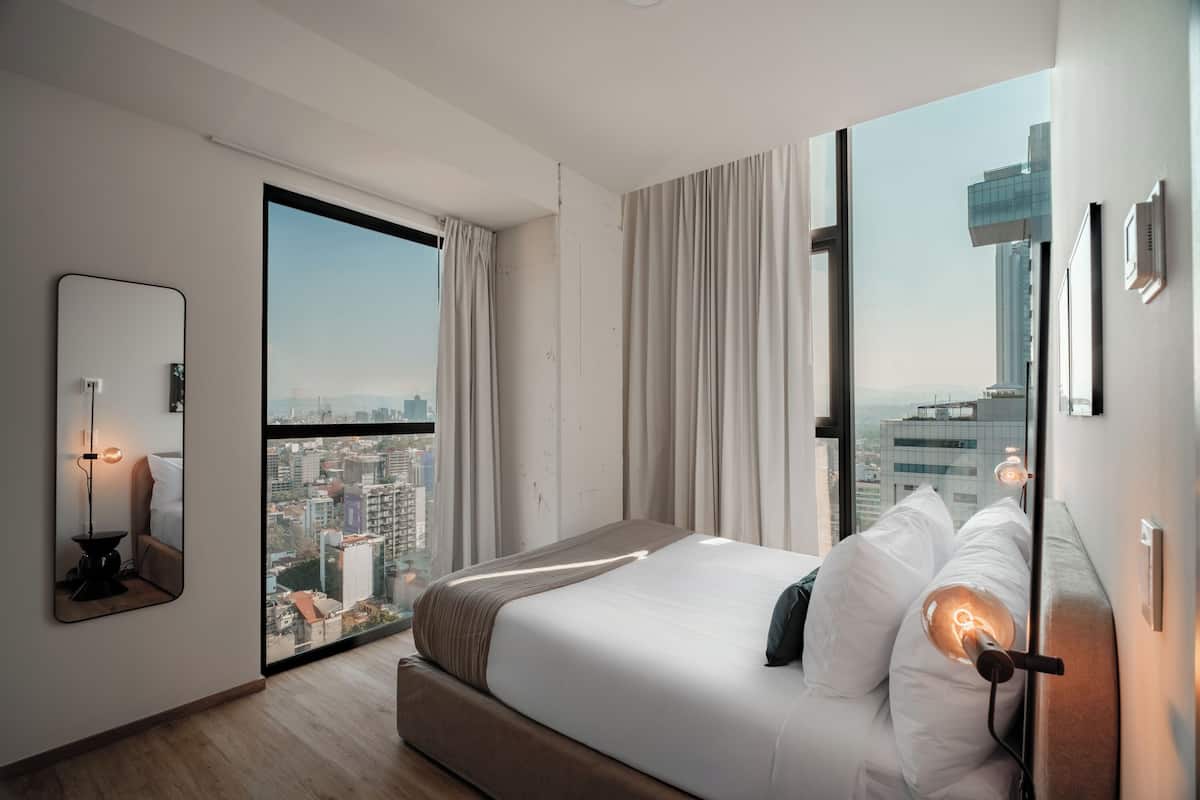 Una habitación de hotel moderna con una cama grande, vista a la ciudad a través de un ventanal de pared a techo, una mesita de noche con lámpara y un espejo.