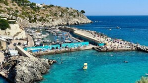 Un pueblo costero con un puerto deportivo, costa rocosa y mar azul cristalino.