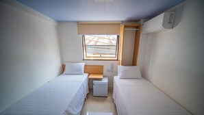 A small, clean room with two single beds, a small table, a window with a view, and an air conditioning unit. - Amazinn Hotel (Belém)