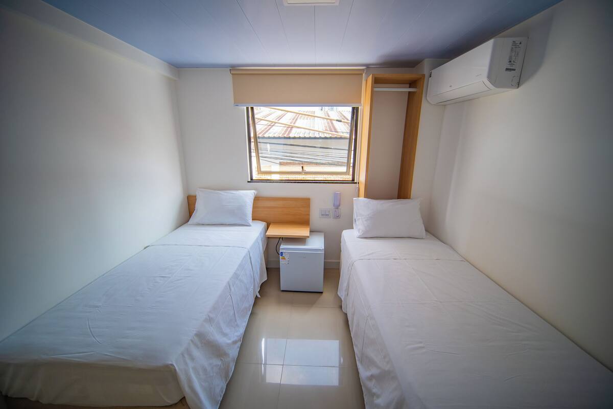 A small, clean room with two single beds, a small table, a window with a view, and an air conditioning unit.