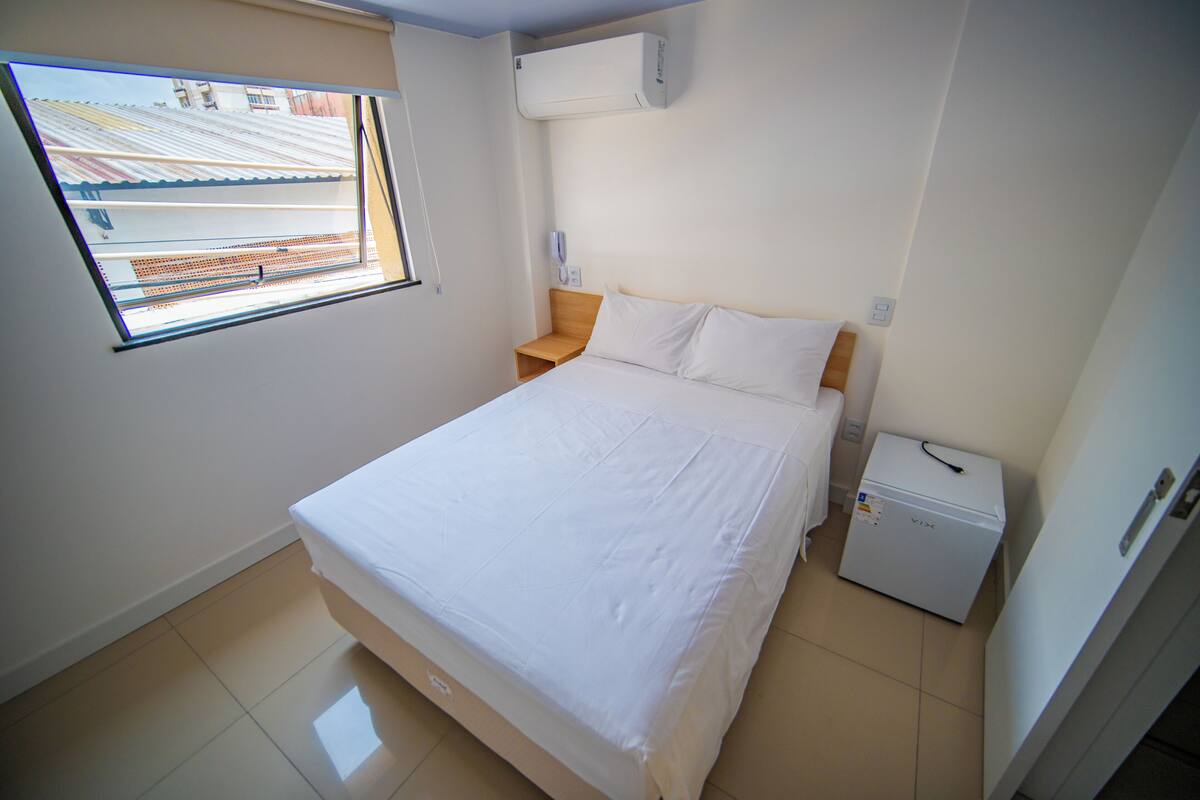 A bedroom with a bed, a nightstand, a small table, an air conditioner, and a window.