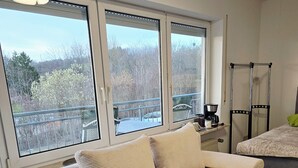 A modern living room with a beige sofa, a coffee table, and a view of trees through large windows. - Apartment near the clinic (Idar-Oberstein)