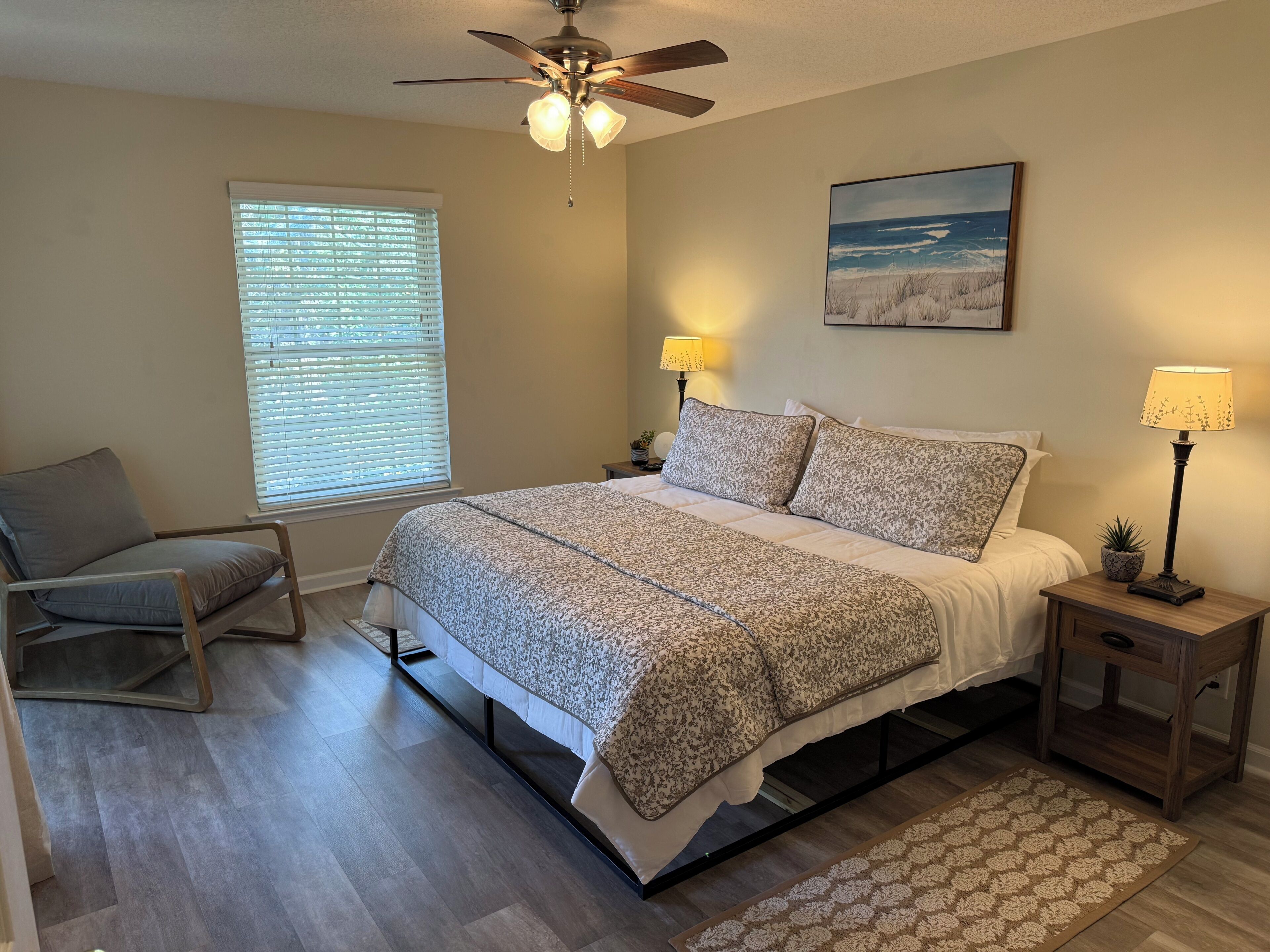 Une chambre à coucher avec un lit, deux tables de chevet avec des lampes, une chaise, un ventilateur de plafond, une fenêtre avec des stores et un tableau accroché au mur.