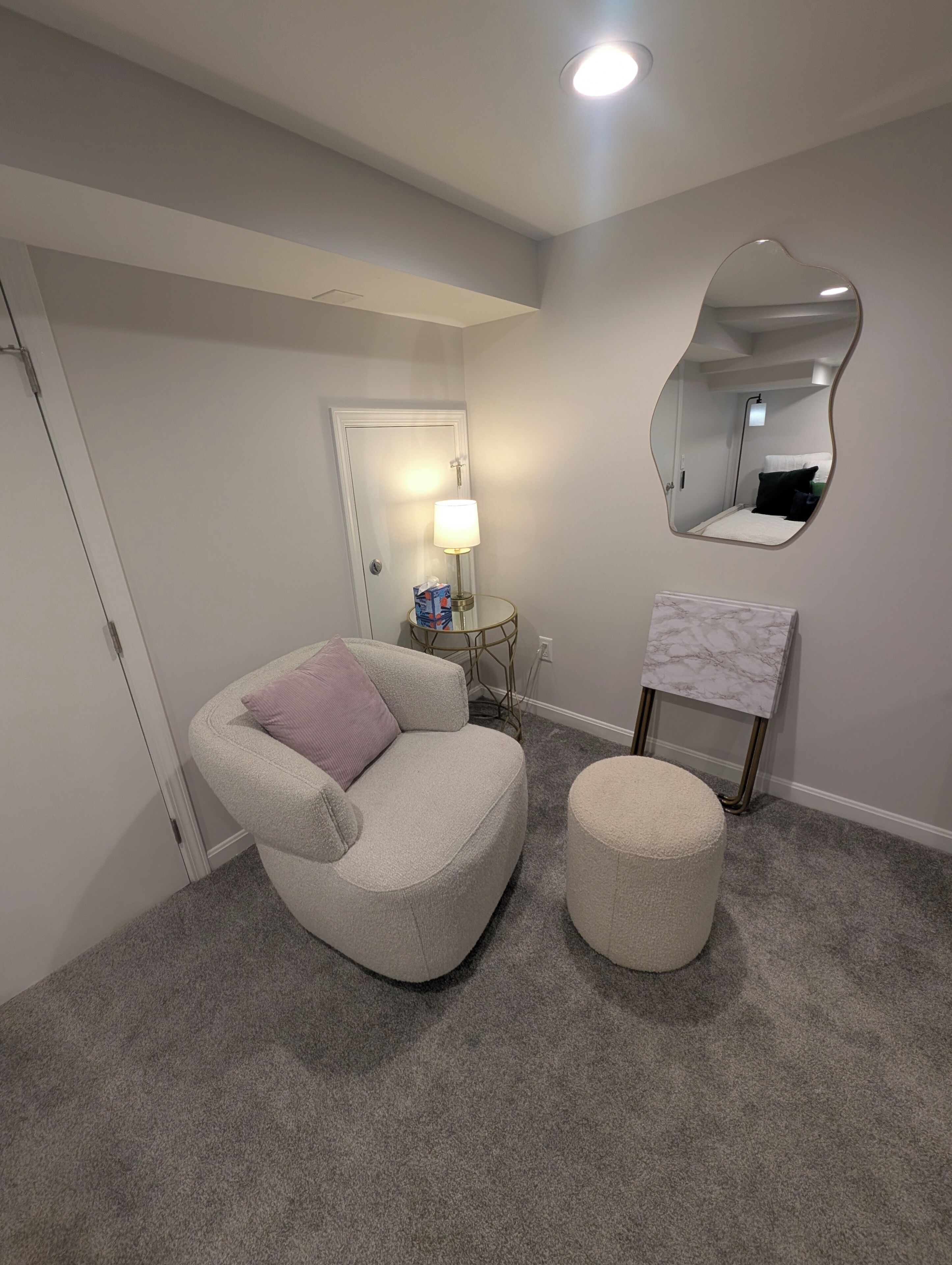 Une pièce avec un tapis gris, un fauteuil gris avec un coussin, un petit pouf rond, une table d’appoint ronde avec une lampe, un miroir au mur et une porte au fond.