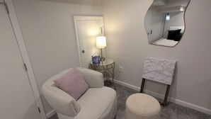 Une pièce avec un tapis gris, un fauteuil gris avec un coussin, un petit pouf rond, une table d’appoint ronde avec une lampe, un miroir au mur et une porte au fond.