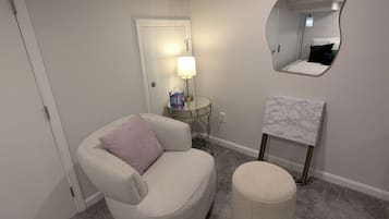 Une pièce avec un tapis gris, un fauteuil gris avec un coussin, un petit pouf rond, une table d’appoint ronde avec une lampe, un miroir au mur et une porte au fond.