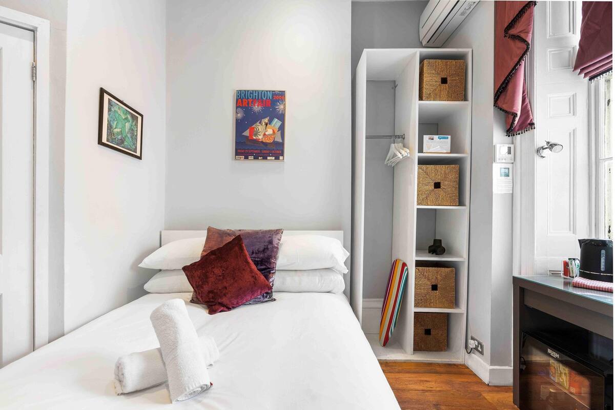 A neatly arranged bedroom with a bed, pillows, a poster, a shelf with storage boxes, and a window with curtains.