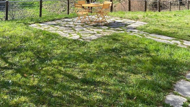 A stone pathway leading to a wooden table and chairs in a grassy area with a mountain in the background.