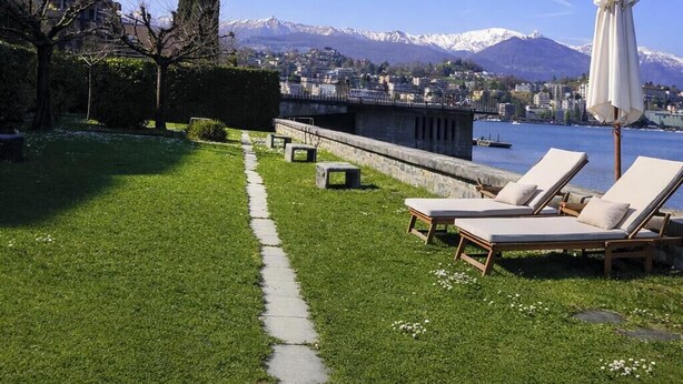Une zone herbeuse avec un sentier menant à un lac, des montagnes se dressant à l’arrière-plan. On y trouve des chaises longues et un parasol.
