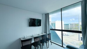 A modern room with a dining table, chairs, a flat-screen TV mounted on the wall, and a large window offering a view of the cityscape.