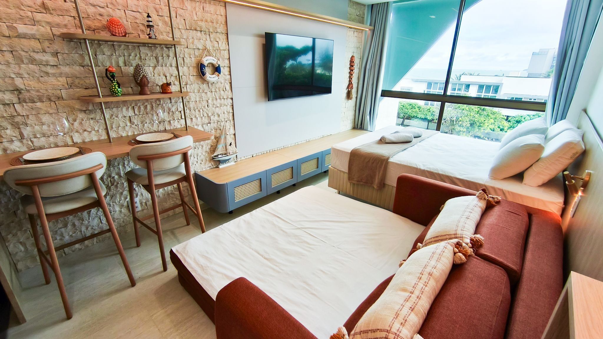 Quarto de hotel moderno com parede de tijolos, televisão de tela plana, cama com roupa de cama vermelha, escrivaninha com cadeira e janela com vista para a cidade.