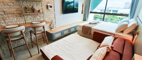 A modern hotel room with a brick wall, a flat-screen TV, a bed with red bedding, a desk with a chair, and a window with a city view.