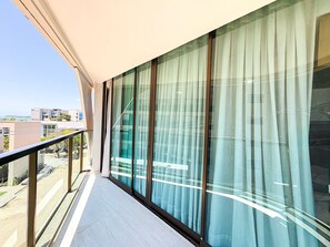 Un balcon doté d’une balustrade en verre, de grandes fenêtres avec des rideaux et une vue sur des immeubles et la verdure.