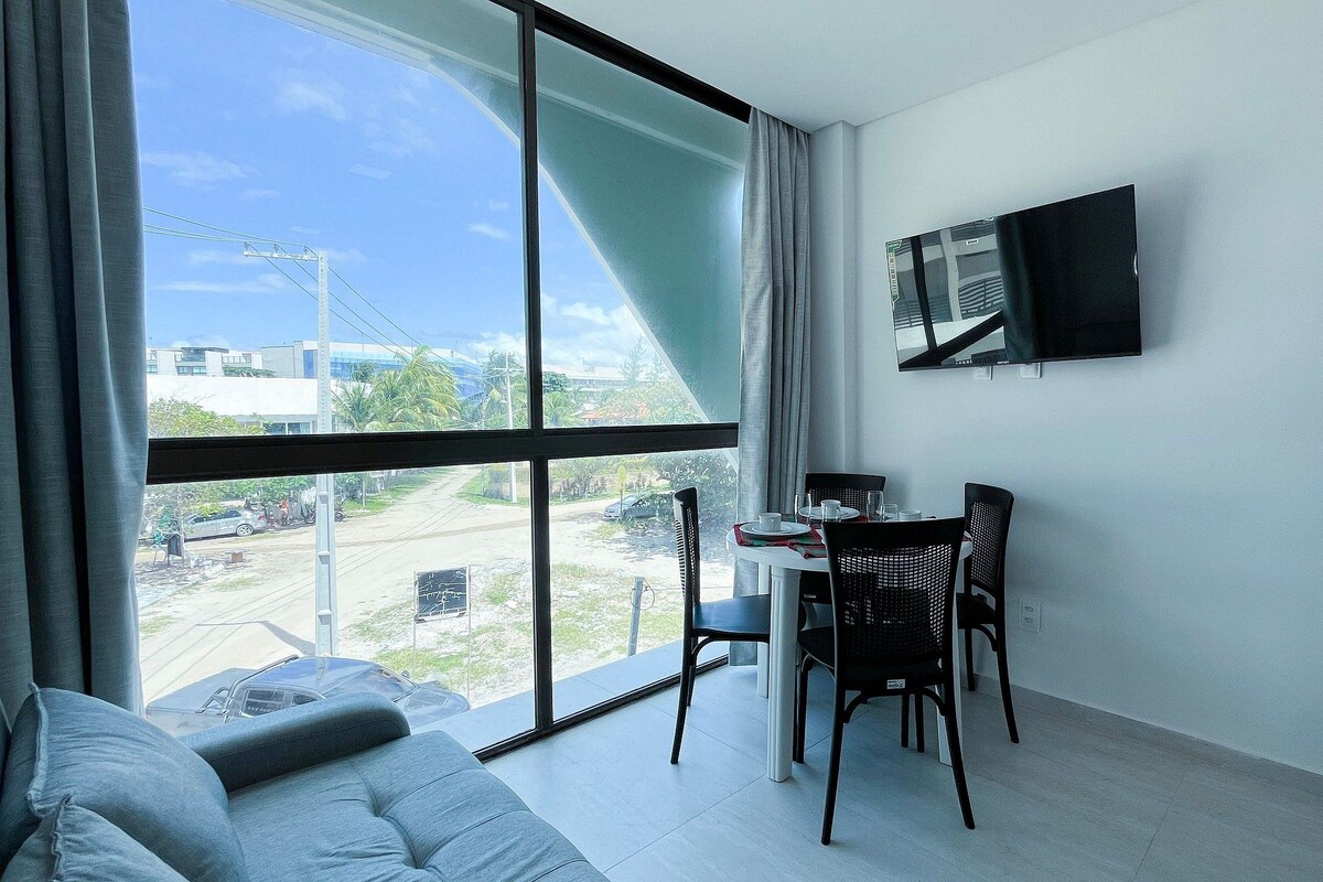 A room with a large window, a dining table with chairs, a flat-screen TV mounted on the wall, and a sofa.