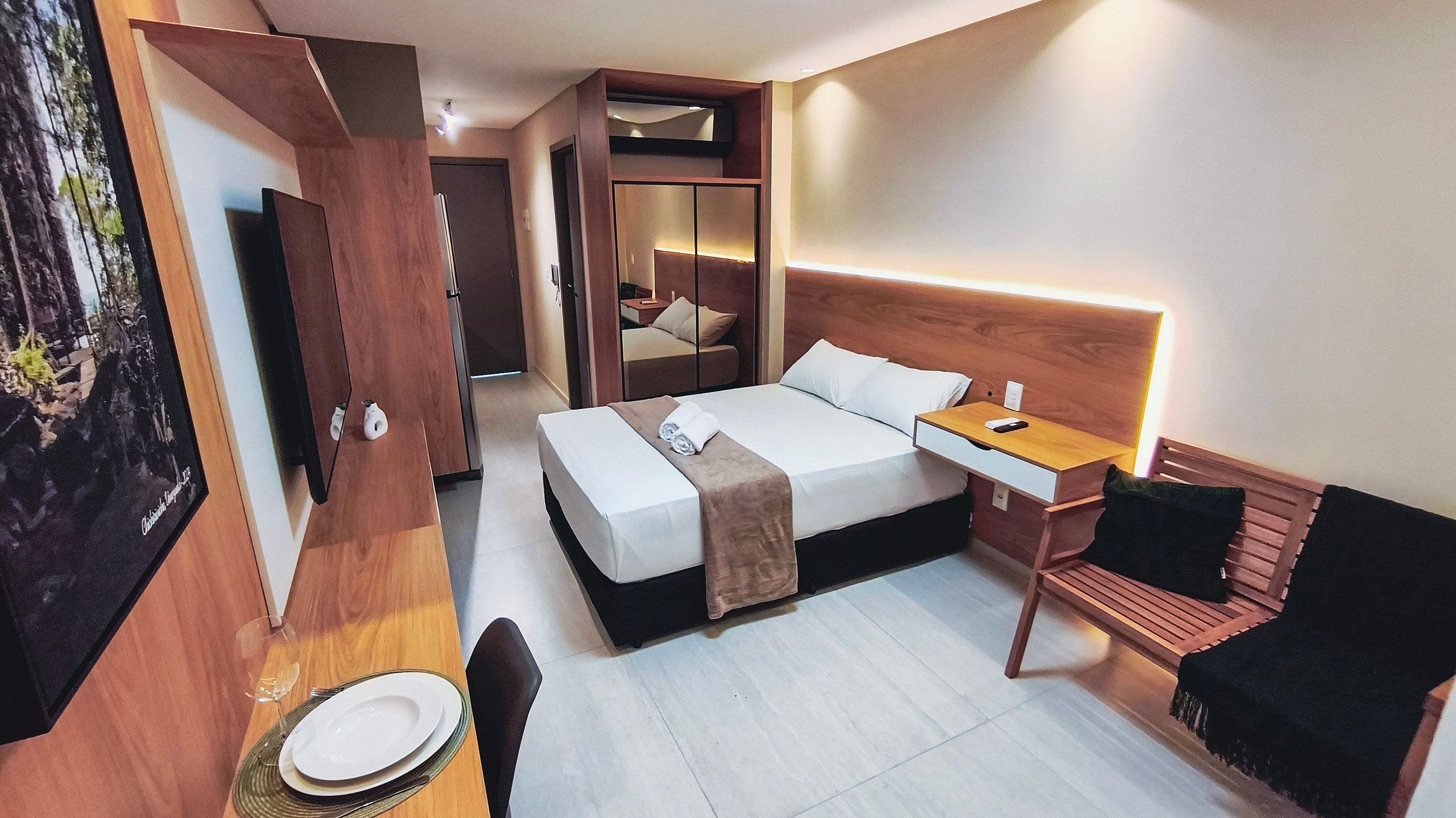 Een hotelkamer met een bed, nachtkastje, stoel en een aan de muur bevestigde televisie.