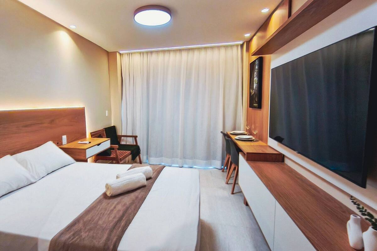 A hotel room with a bed, bedside tables, a desk, a chair, a TV, and a window with curtains.