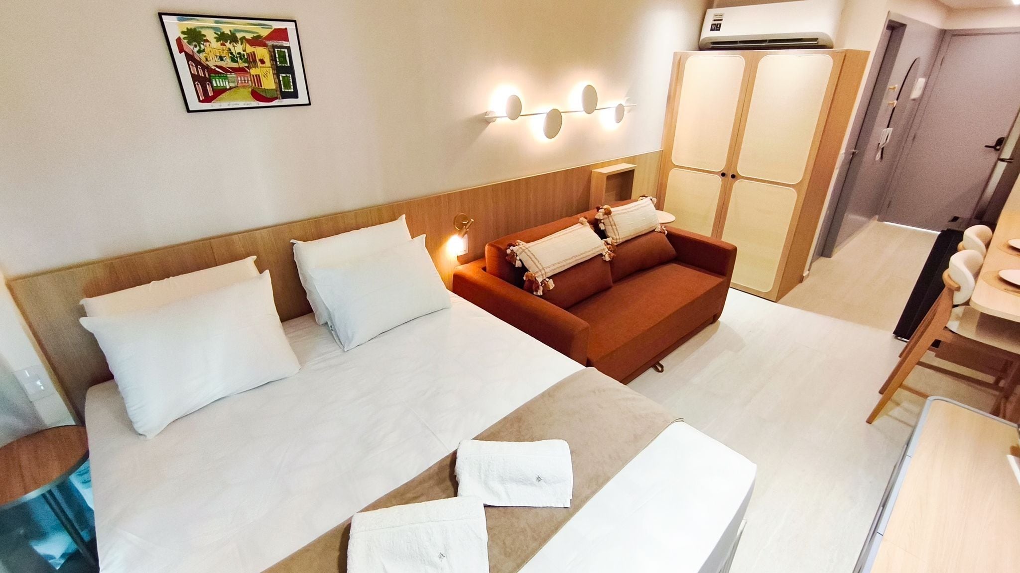 A hotel room with a bed, a sofa, a table, and a wall-mounted air conditioning unit.