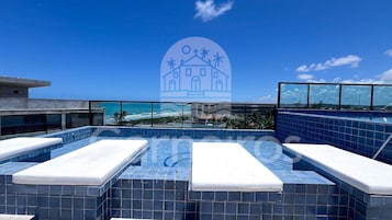 Una piscina en la azotea con baldosas azules, vista al océano y un edificio con palmeras al fondo.