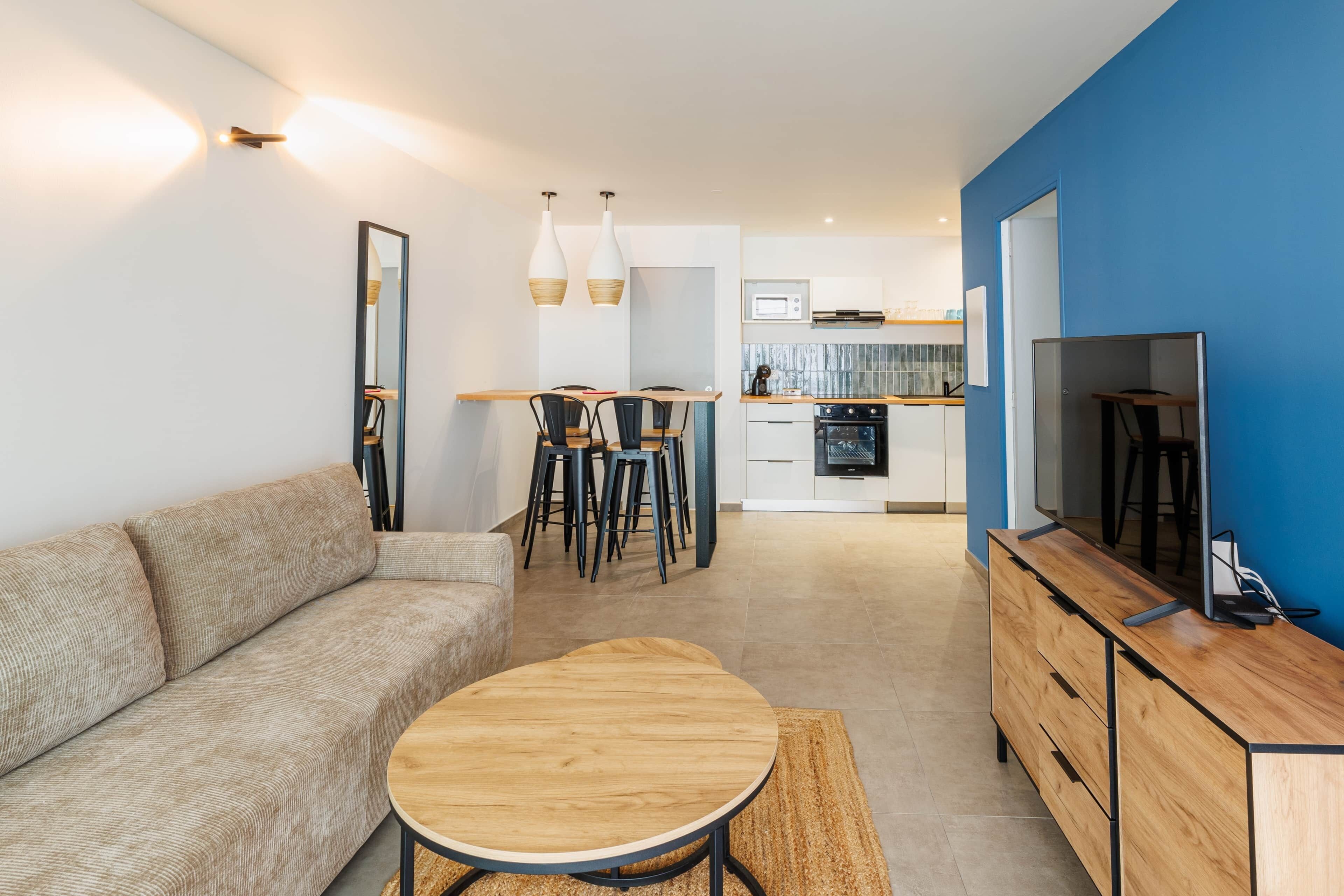 Uma sala de estar moderna com sofá, mesa de centro de madeira e TV em um móvel de madeira. Há um espaço de jantar com cadeiras e uma cozinha ao fundo.