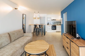 A modern living room with a sofa, wooden coffee table, and a TV on a wooden cabinet. There is a dining area with chairs and a kitchen in the background. - Flat with sea view (Saint Pierre)