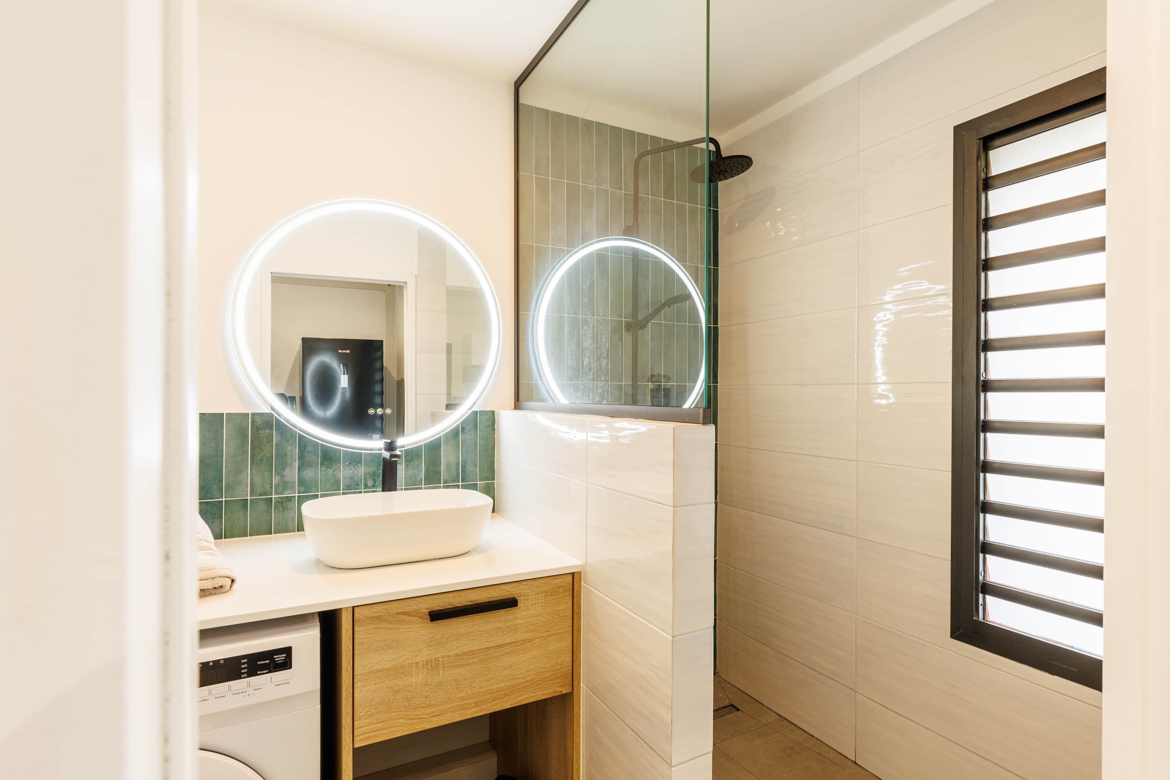 Une salle de bain avec un lavabo blanc, deux miroirs ronds avec éclairage intégré, un meuble en bois et une fenêtre avec des stores.