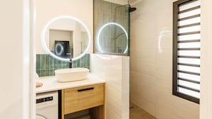 Un bany amb un lavabo blanc, dos miralls rodons amb il·luminació, un armari de fusta i una finestra amb persianes.