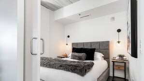 A modern bedroom with a large bed, two bedside tables with lamps, a grey headboard, and a small side table with a plant. - Modern High-Rise Condo | 2 Bed | 1 Bath | (Winnipeg)