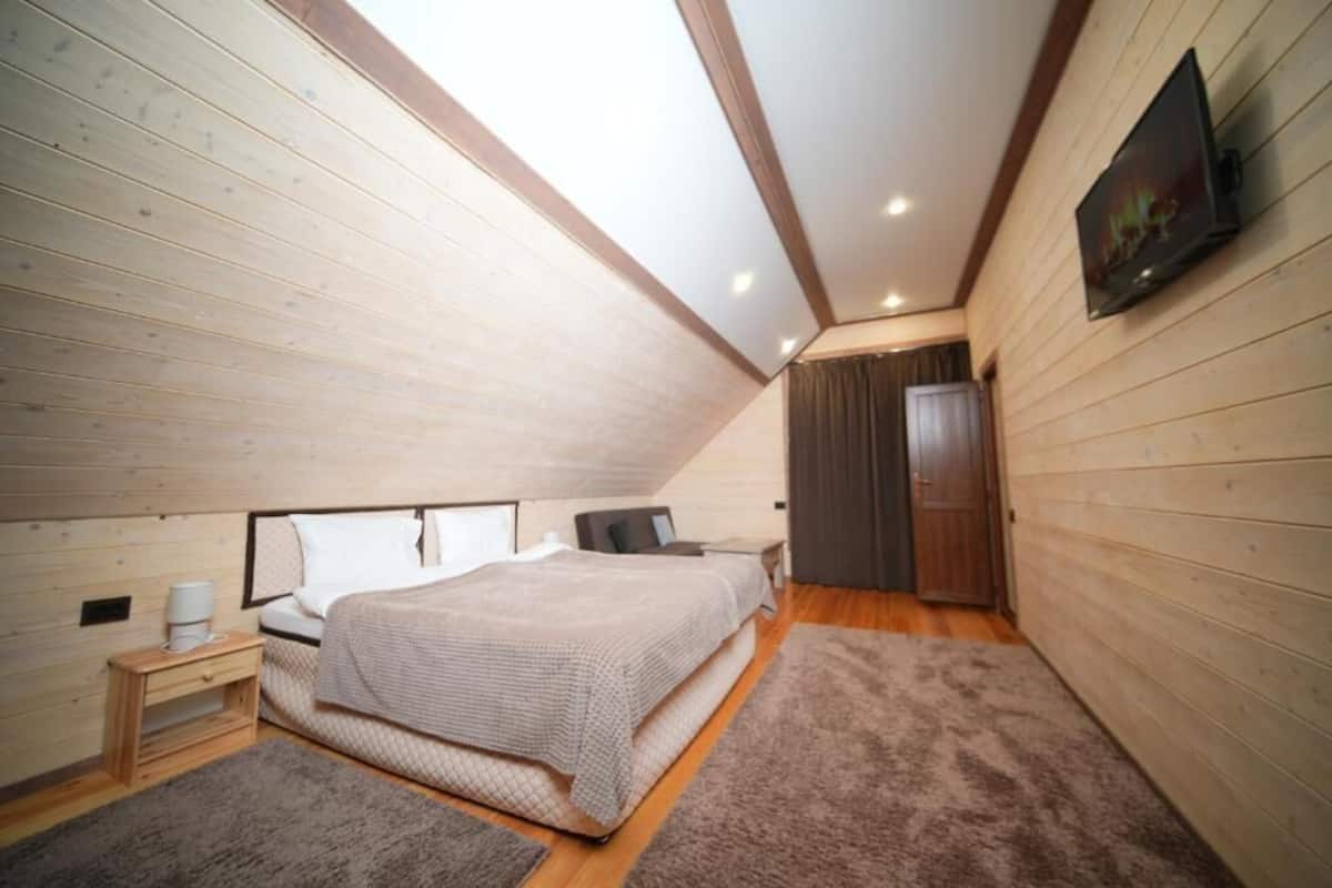 A bedroom with a sloped ceiling, wooden walls, a bed, a nightstand, a TV, and a door.