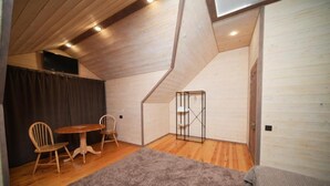 A room with a sloped ceiling, a wooden floor, a round table with chairs, a grey carpet, and a door with a metal gate. - Maru house (Dilijan)