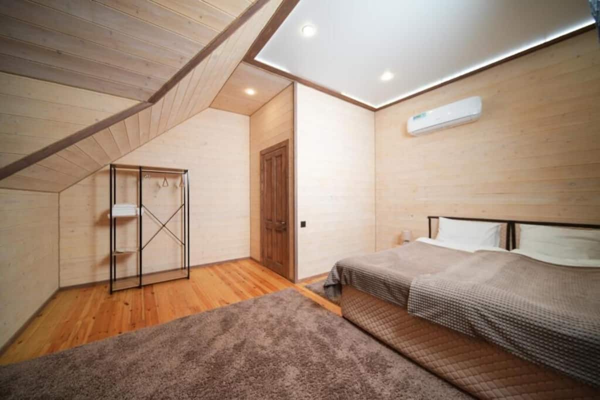 A bedroom with a sloped ceiling, wooden walls, a bed, a small bedside table, and a wall-mounted air conditioner.