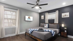 A bedroom with a bed, a ceiling fan, a wall-mounted TV, and a nightstand with a lamp. - 5 Bedrooms w. TVs in every room & Garage Parking (Overland Park)