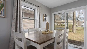 A dining area with a wooden table, white chairs, a window with blinds, and a view of a residential area. - 5 Bedrooms w. TVs in every room & Garage Parking (Overland Park)